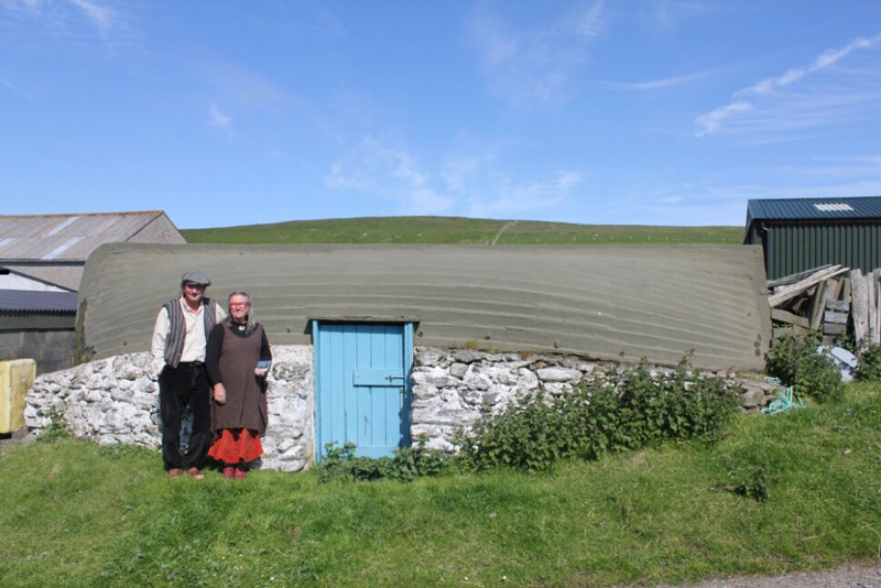 Photo de Solange et Yves Dussin, intervenants lors de la conférence « Les coques de bateaux retournées des îles Shetland », 
						posent devant une chaloupe retournée servant de toiture sur un abri de jardin.