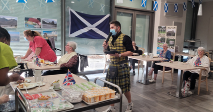 Photo of residents of the EHPAD seated, Bastien Vieuville, animator at the EHPAD 
					is at the microphone, he wears a traditional Scottish outfit for the occasion: a vest and a yellow tartan kilt as well as a 
					sporran (bag) at the belt. The room is also decorated with Scottish flags and pictures of Scotland