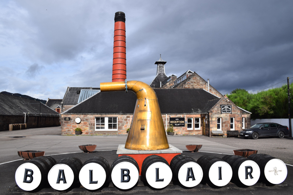 The photo shows the Balblair distillery, located north of Inverness, in the background, with its chimney 
						and its typical roof, with in the foreground a row of whisky barrels, each bearing a letter forming the word Balblair, with a copper 
						still just behind (photo Pierre Delignière).