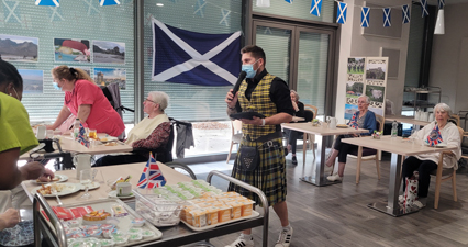 Photo de résidents de l'EHPAD attablés, Bastien Vieuville, animateur à l’EHPAD est au 
					micro, il porte une tenue traditionnelle écossaise pour l'occasion : un gilet et un kilt en tartan jaune ainsi qu'un sporran (sacoche)
					à la ceinture. La pièce est également décorée avec des drapeaux écossais et des photos de l'Ecosse