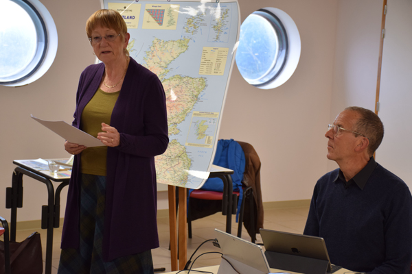 Mary Chapman, standing left, in front of a map of Scotland, and Andrew Pears, sitting right, 
					recount their sailing trip around Scotland.